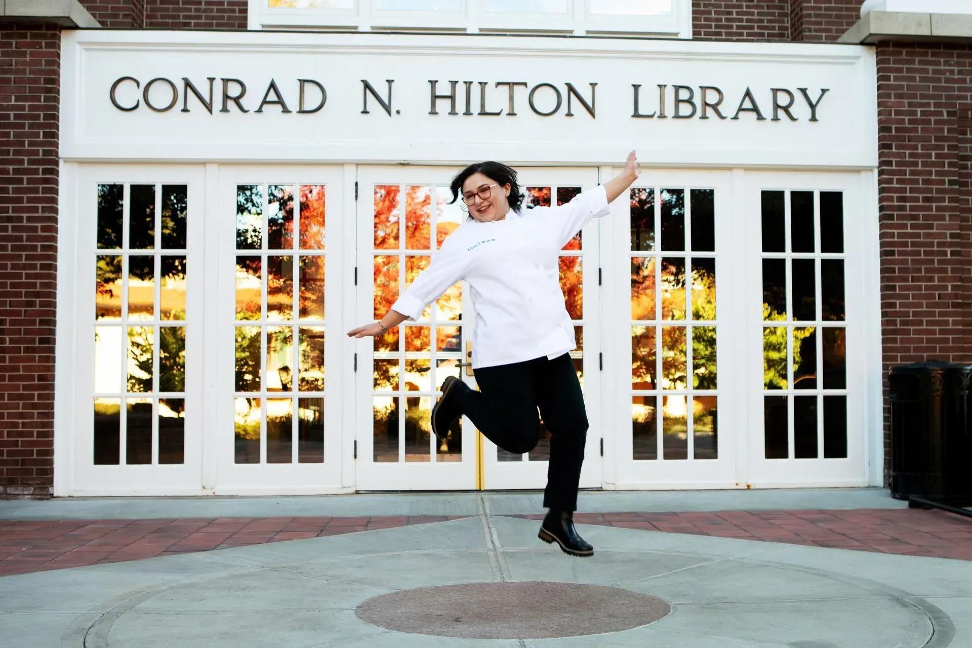 Person jumps for joy in front of the Conrad N. Hilton Library. White shirt, black pants, brick building, fall foliage.