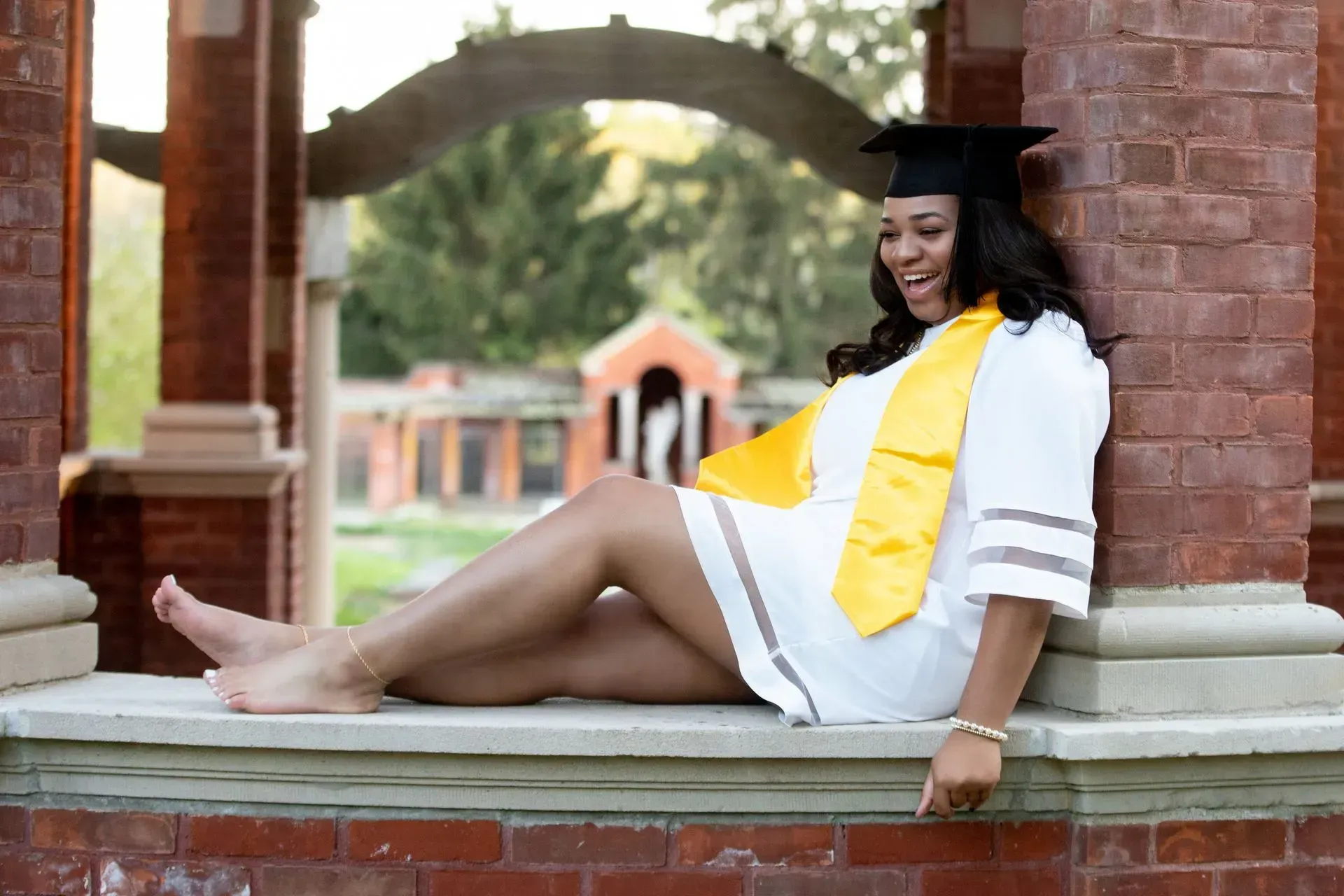 Graduation student in white dress and gold sash, leaning on a brick ledge, laughing.