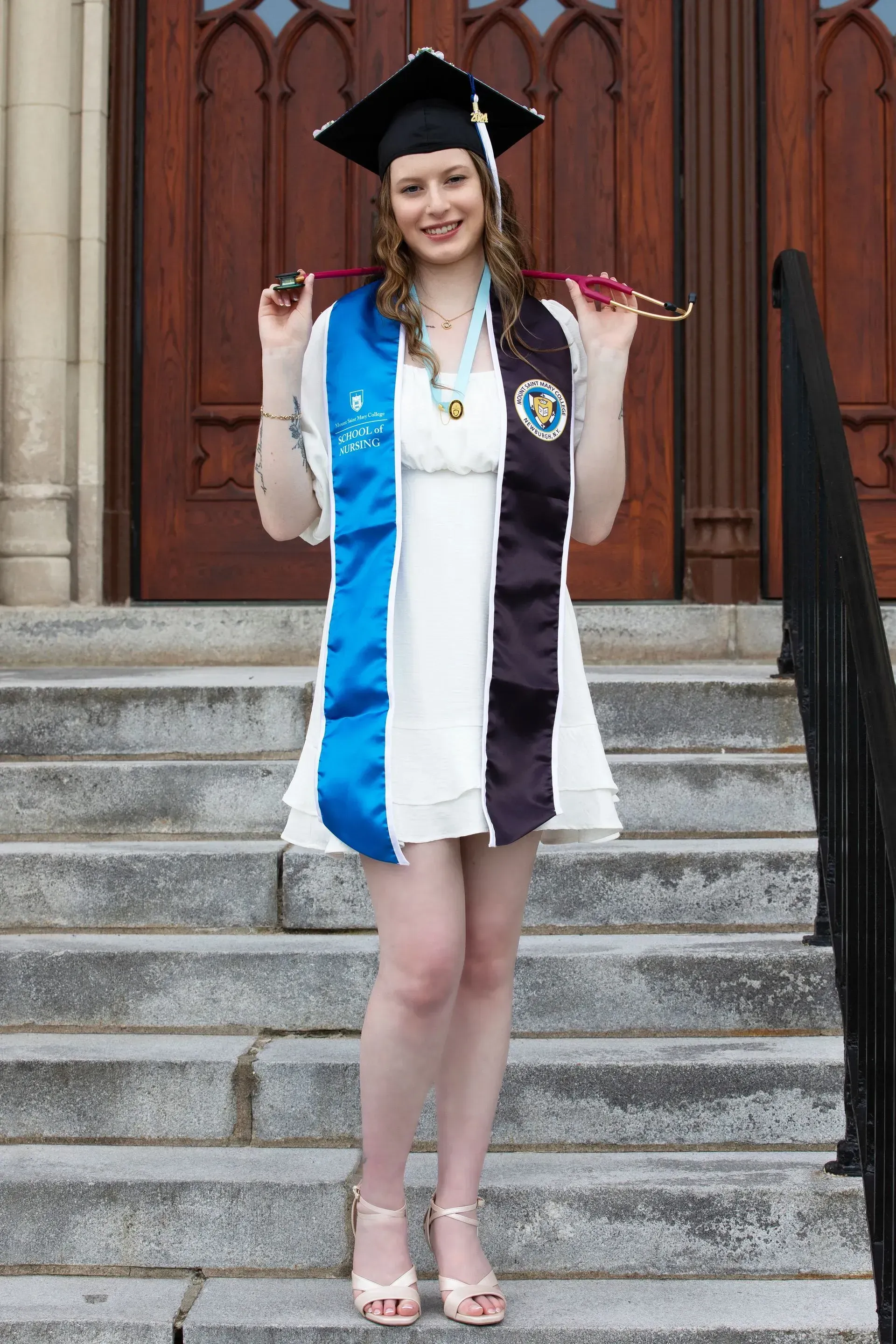 Woman in graduation attire stands on steps, holding stethoscope. Blue and black sashes, white dress, black cap.