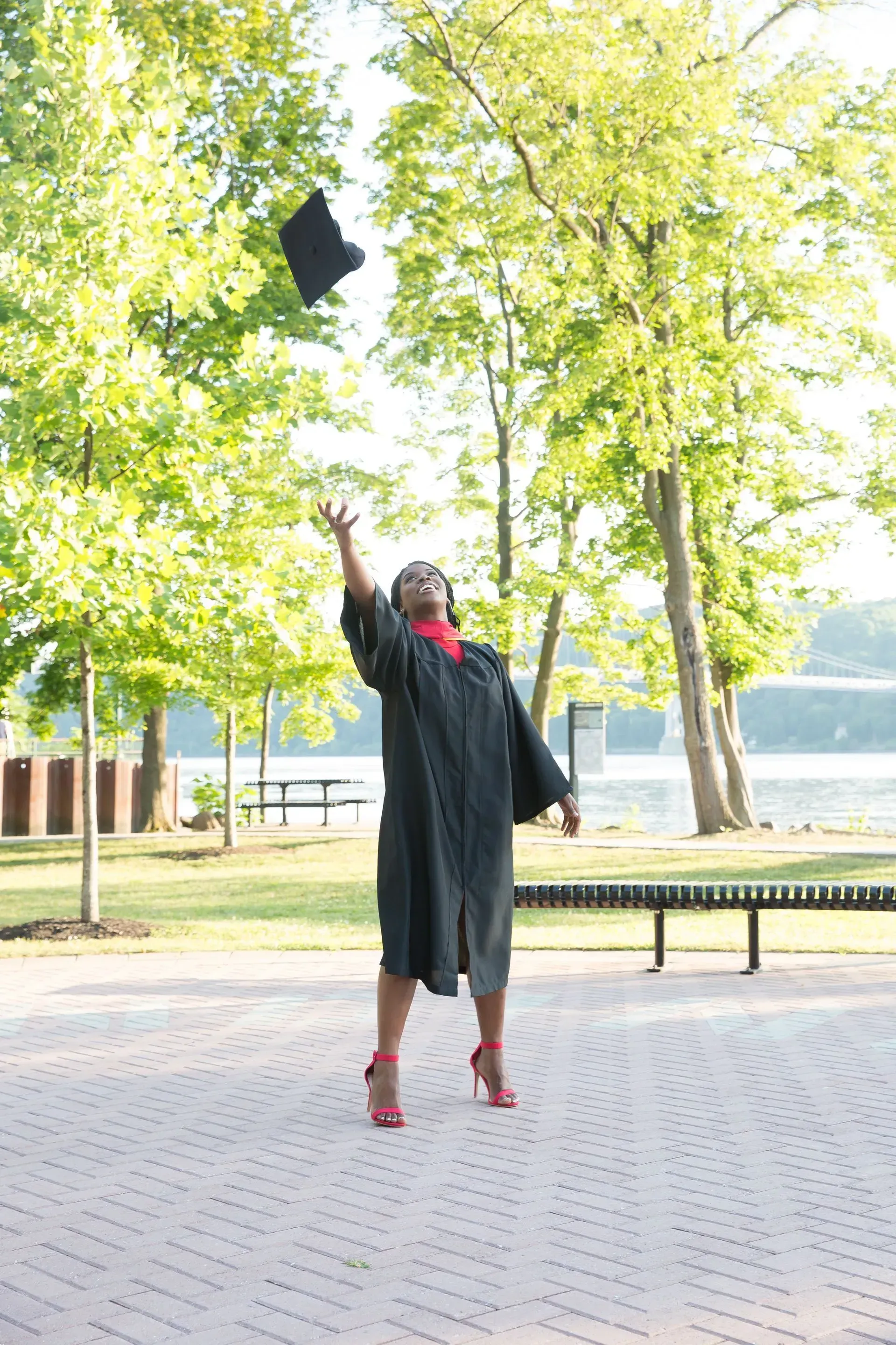 Person in graduation gown throws cap in air, red heels, park setting with trees and water.