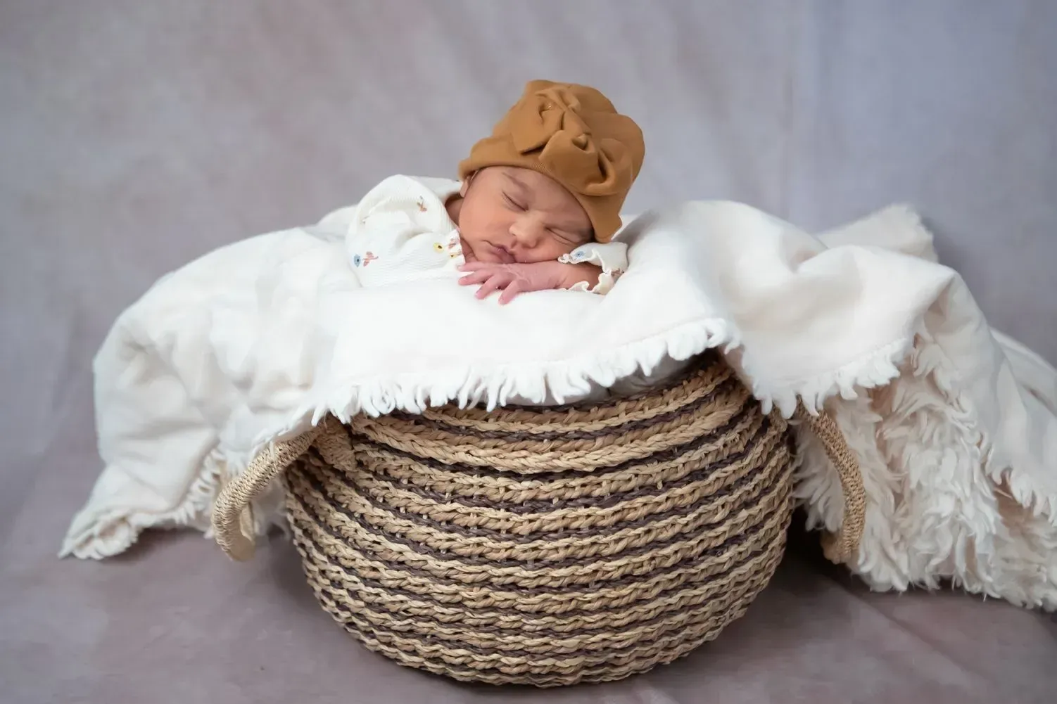 Newborn baby sleeping in a woven basket, wearing a brown hat and covered with a white blanket on a neutral backdrop.