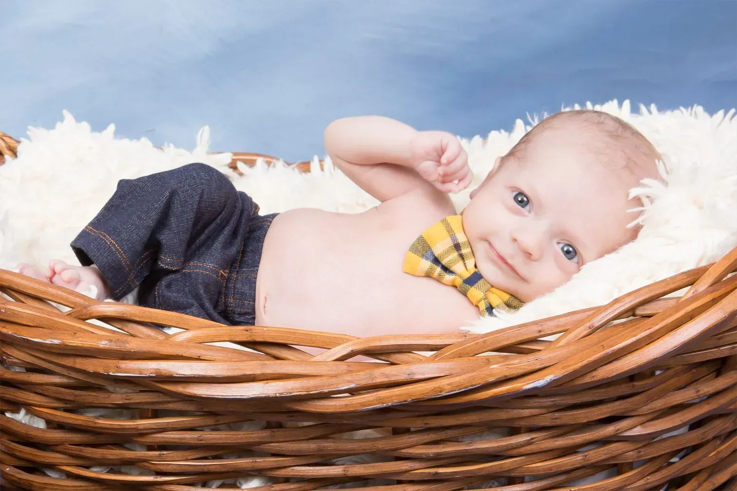 Baby in a basket with a yellow bowtie and jeans, looking at the viewer.