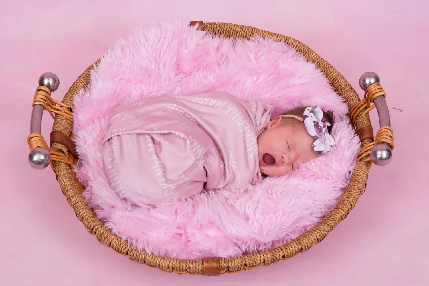 Newborn baby swaddled in pink, asleep in a fluffy pink-lined basket with a floral headband.