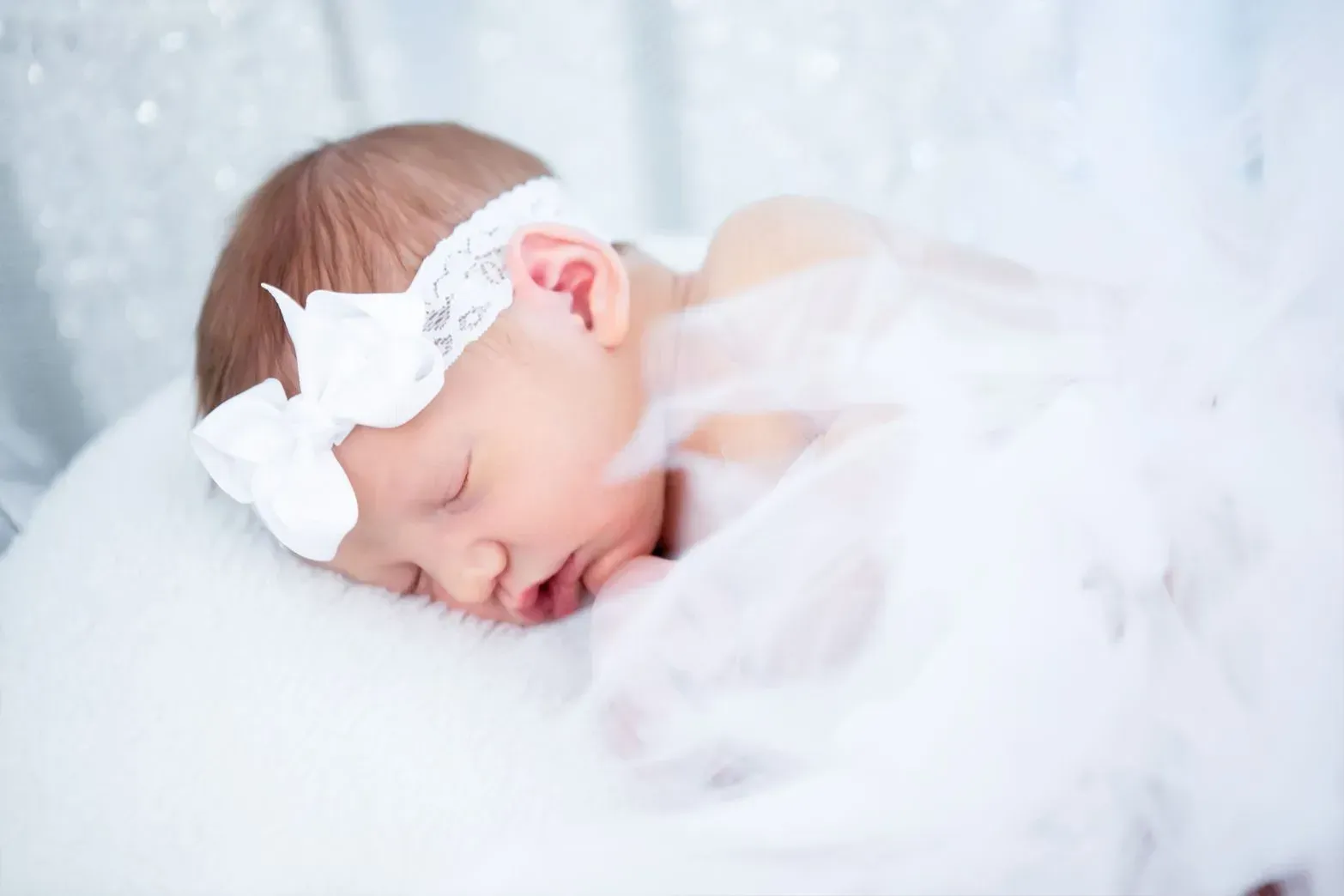 Sleeping newborn wearing a white lace headband and tulle wrap on a white cushion.