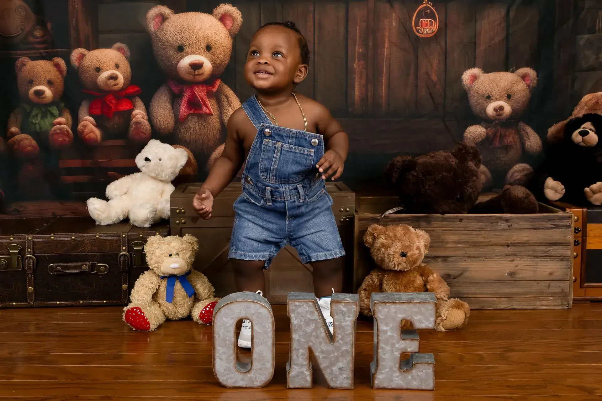 Child in denim overalls smiles, surrounded by teddy bears. Wooden letters spell 