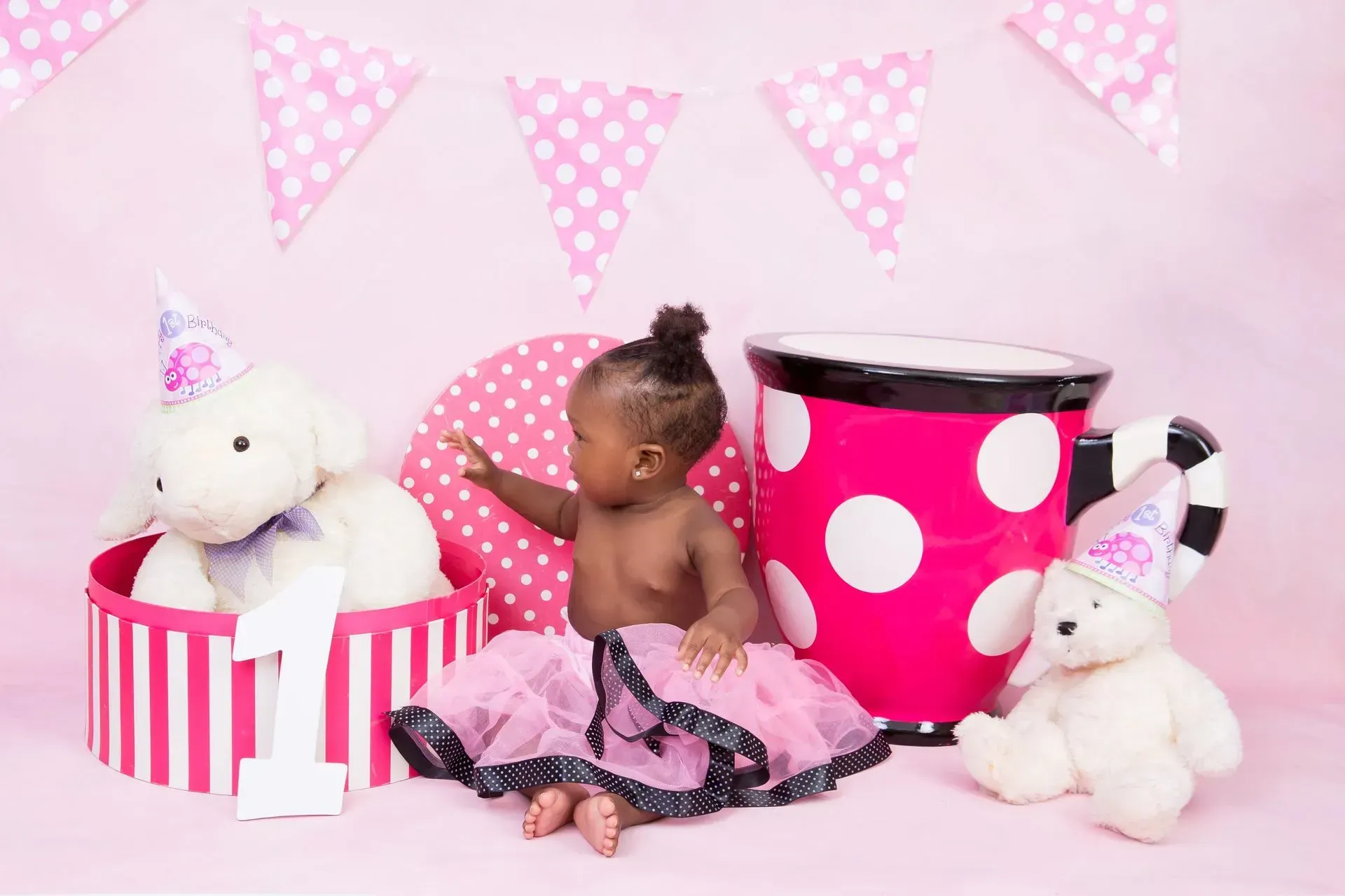 A baby in a pink tutu sits near a teacup, toys, and decorations for a first birthday.
