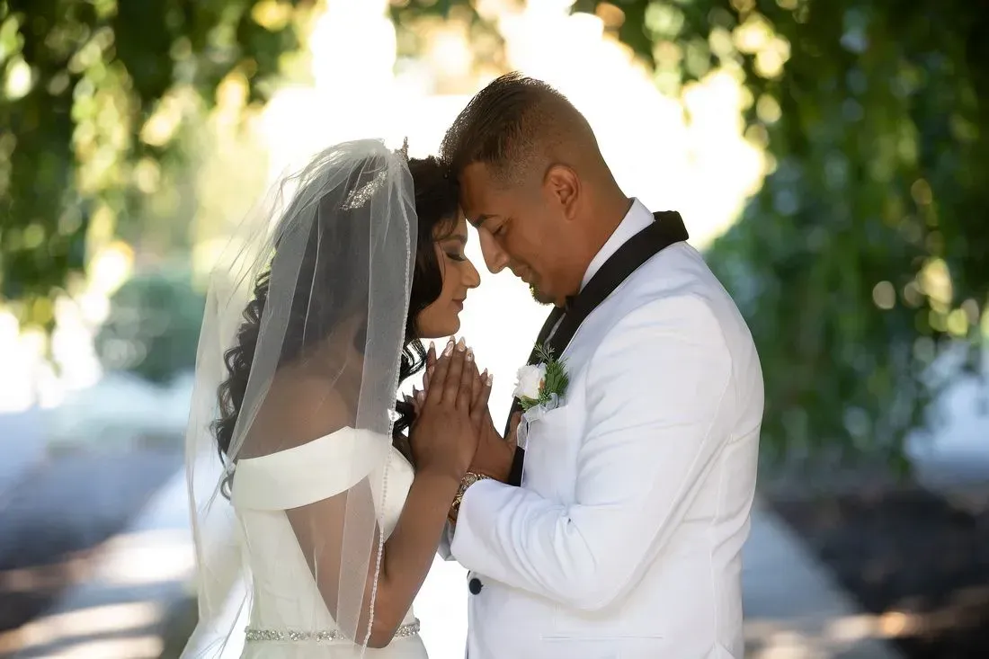 Bride and groom holding hands, foreheads touching, on a sunny day. She wears a veil, he wears a white suit.