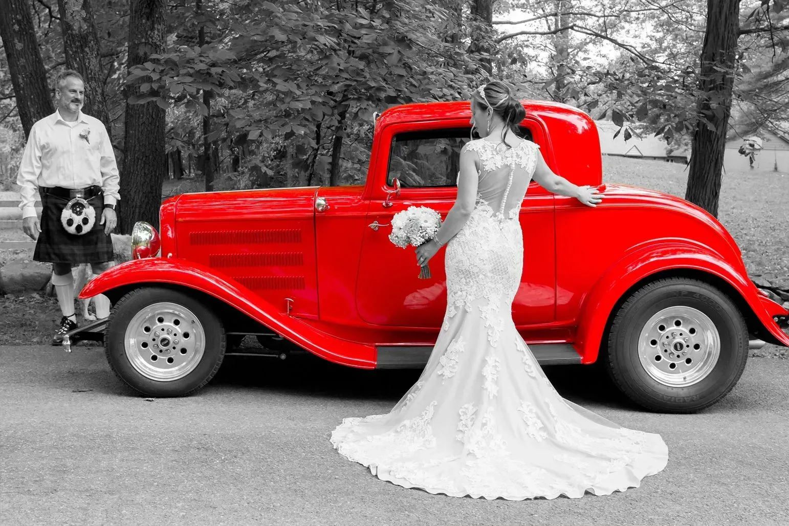 Bride in lace dress beside a bright red classic car. Man in kilt stands nearby.