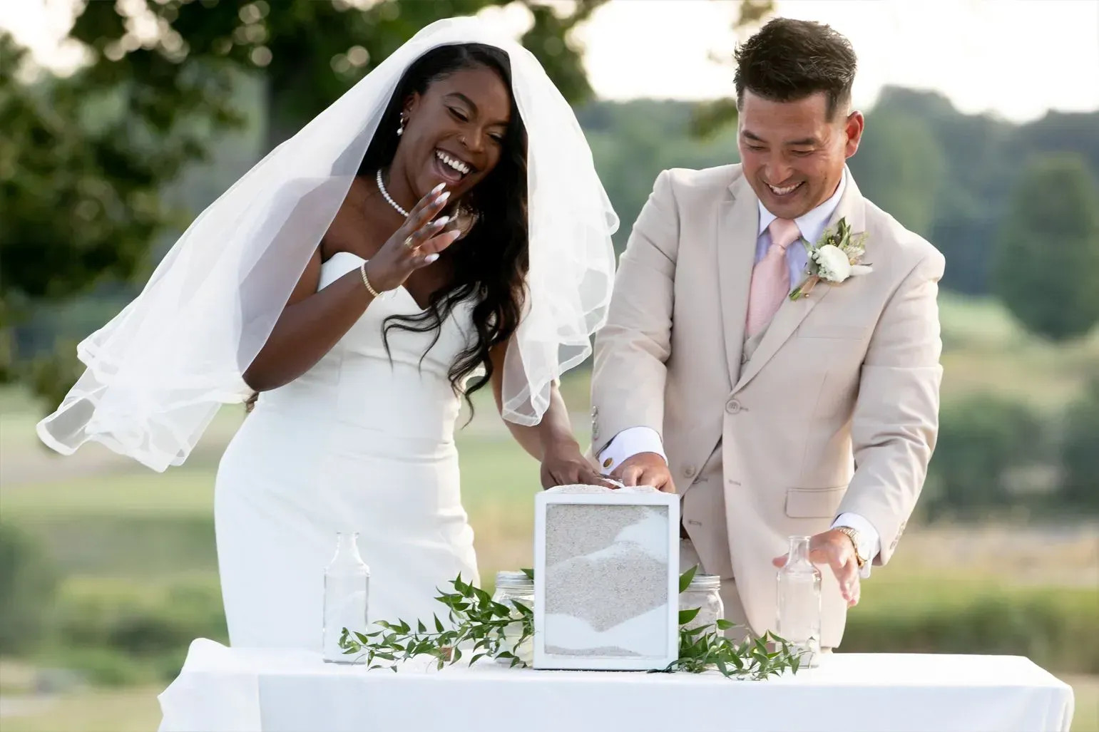 Couple at wedding ceremony pouring sand into a frame, smiling. Outdoor setting.