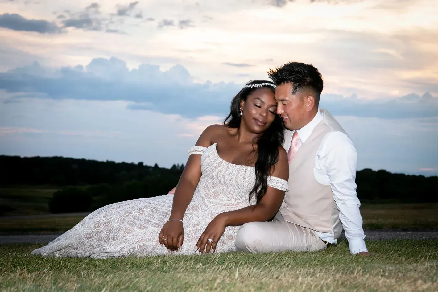 Bride and groom sit on grass, embracing, against a sunset sky.