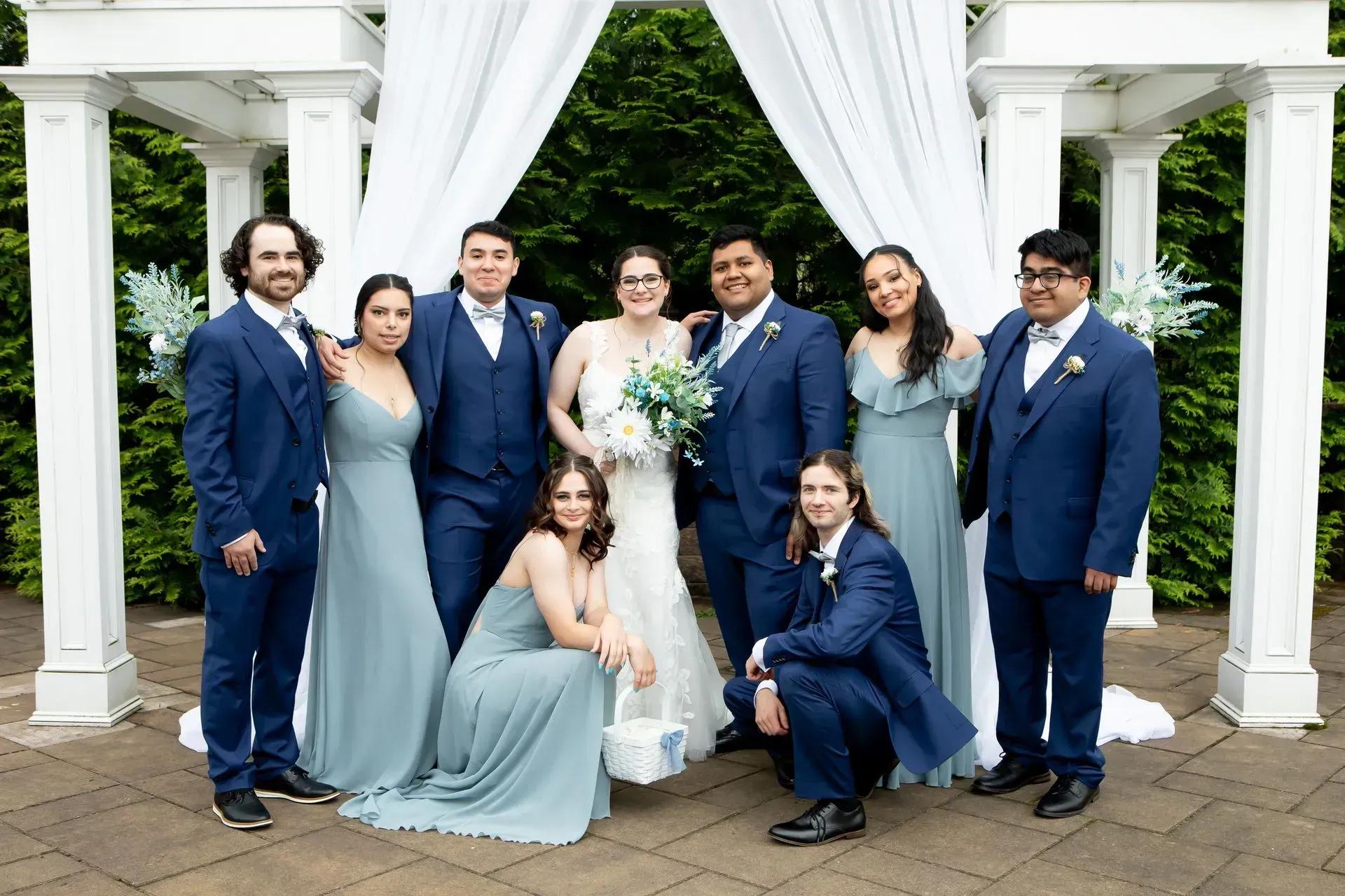 Wedding group portrait in front of white arbor with bride, groom, and wedding party in blue.
