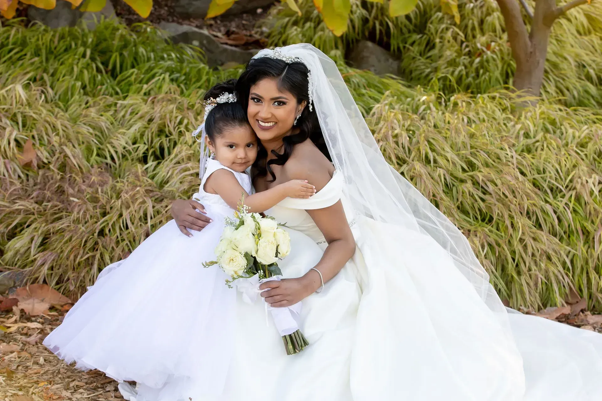 Bride in white gown embraces flower girl in white dress, holding bouquet.