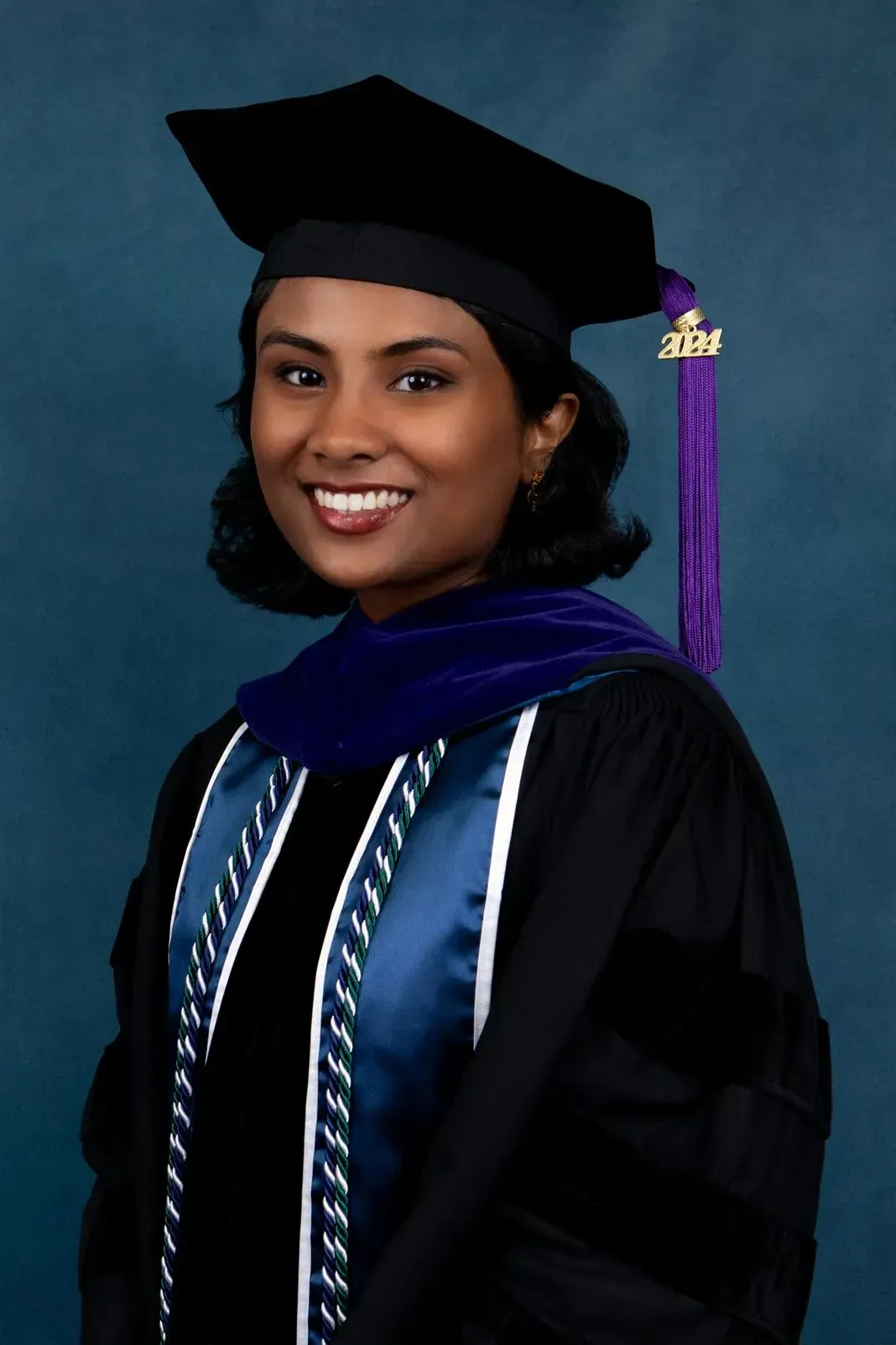 Woman in graduation attire, smiling, with purple tassel, blue and black robes against a dark blue background.