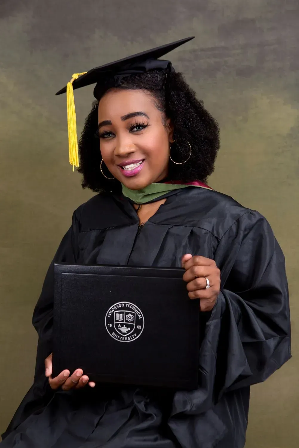 Person in graduation gown holding diploma, smiling, against a blurred backdrop.