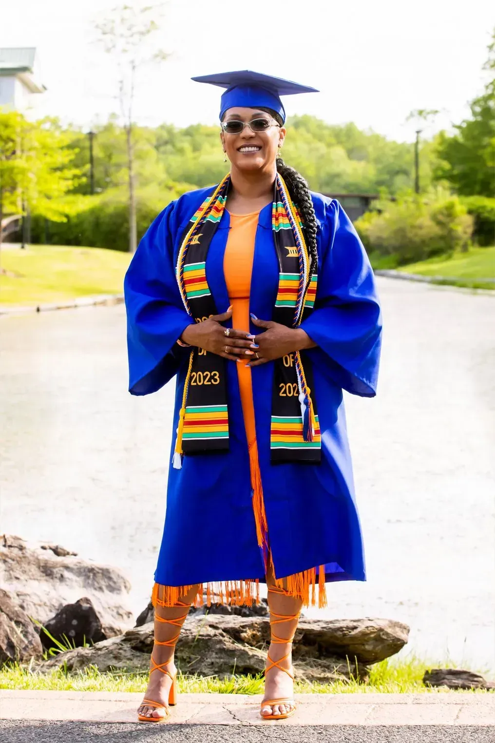 Woman in blue graduation gown and cap, orange dress, holding a sash, standing outside.