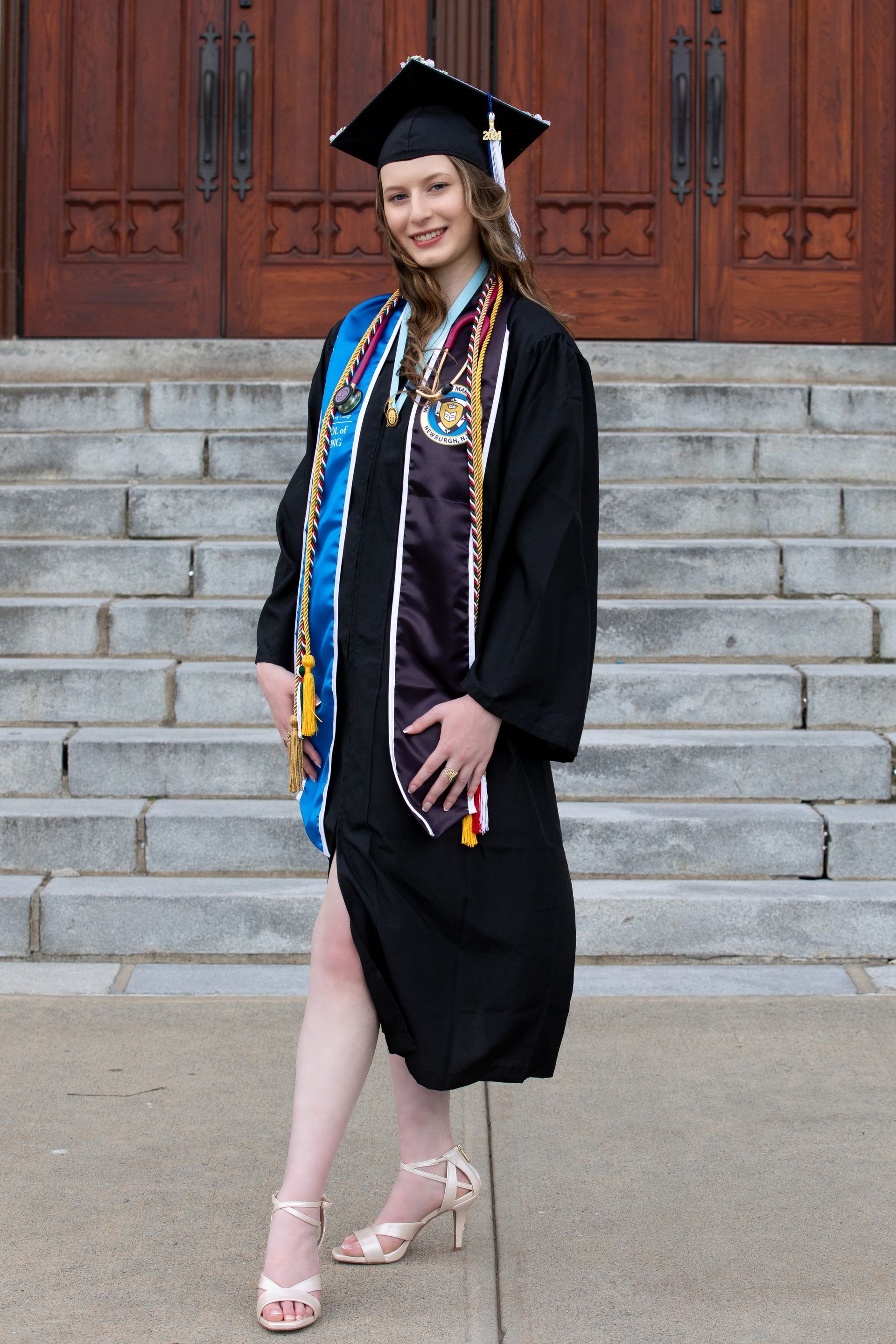 A person in a graduation gown, cap, and stoles poses smiling on outdoor stone steps in front of wooden double doors.