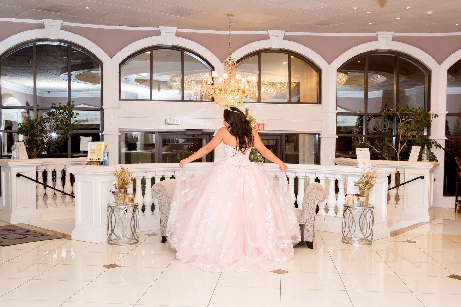 A person in a voluminous light pink gown stands facing away from the camera in a grand, arched, white-tiled lobby. 