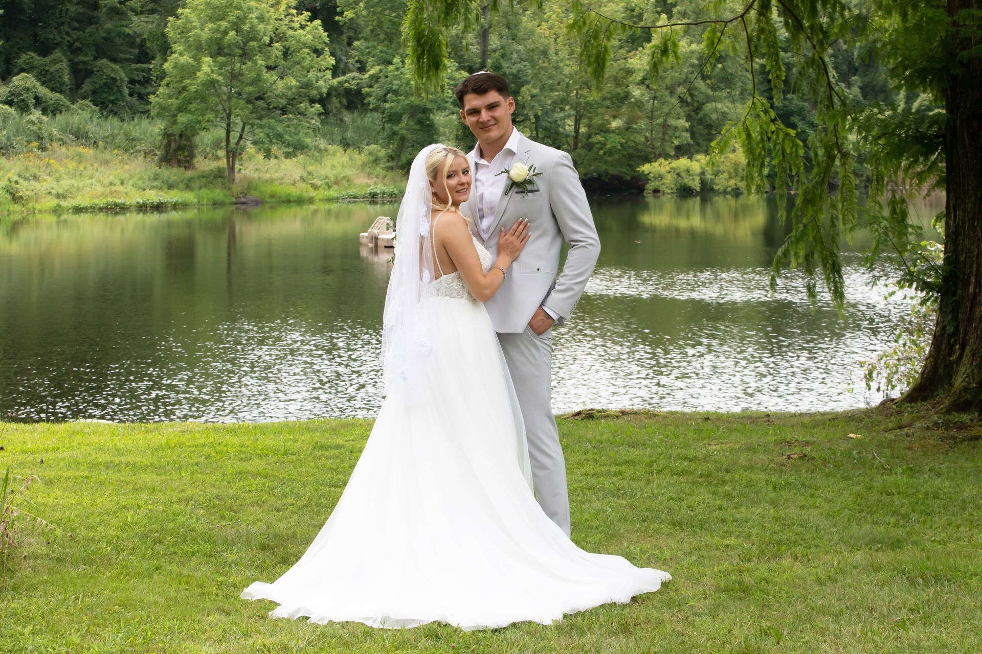 A couple in wedding attire poses together in a green park beside a calm lake.