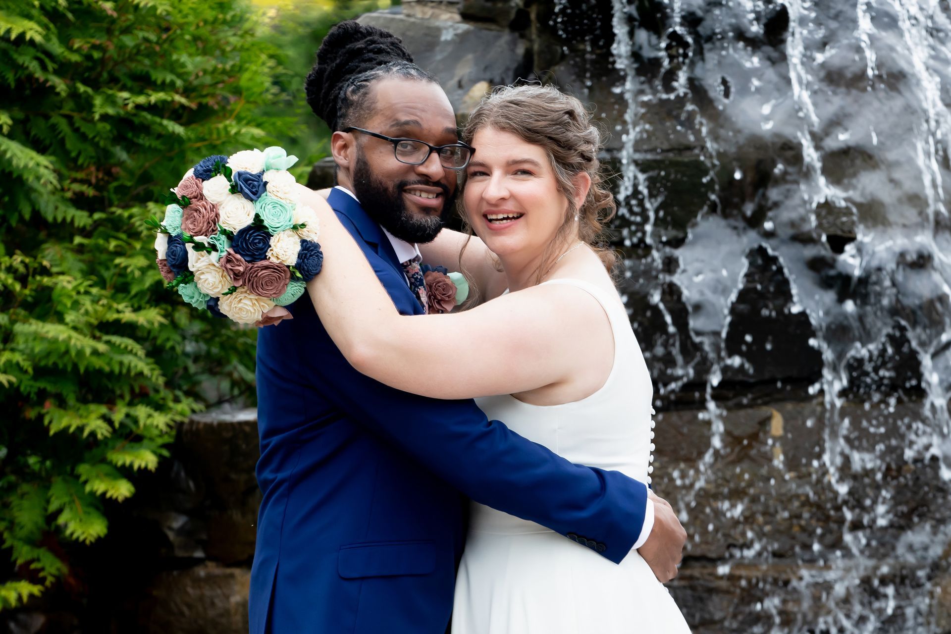A bride and groom in wedding attire embrace in front of a waterfall, the bride holding a colorful bouquet.