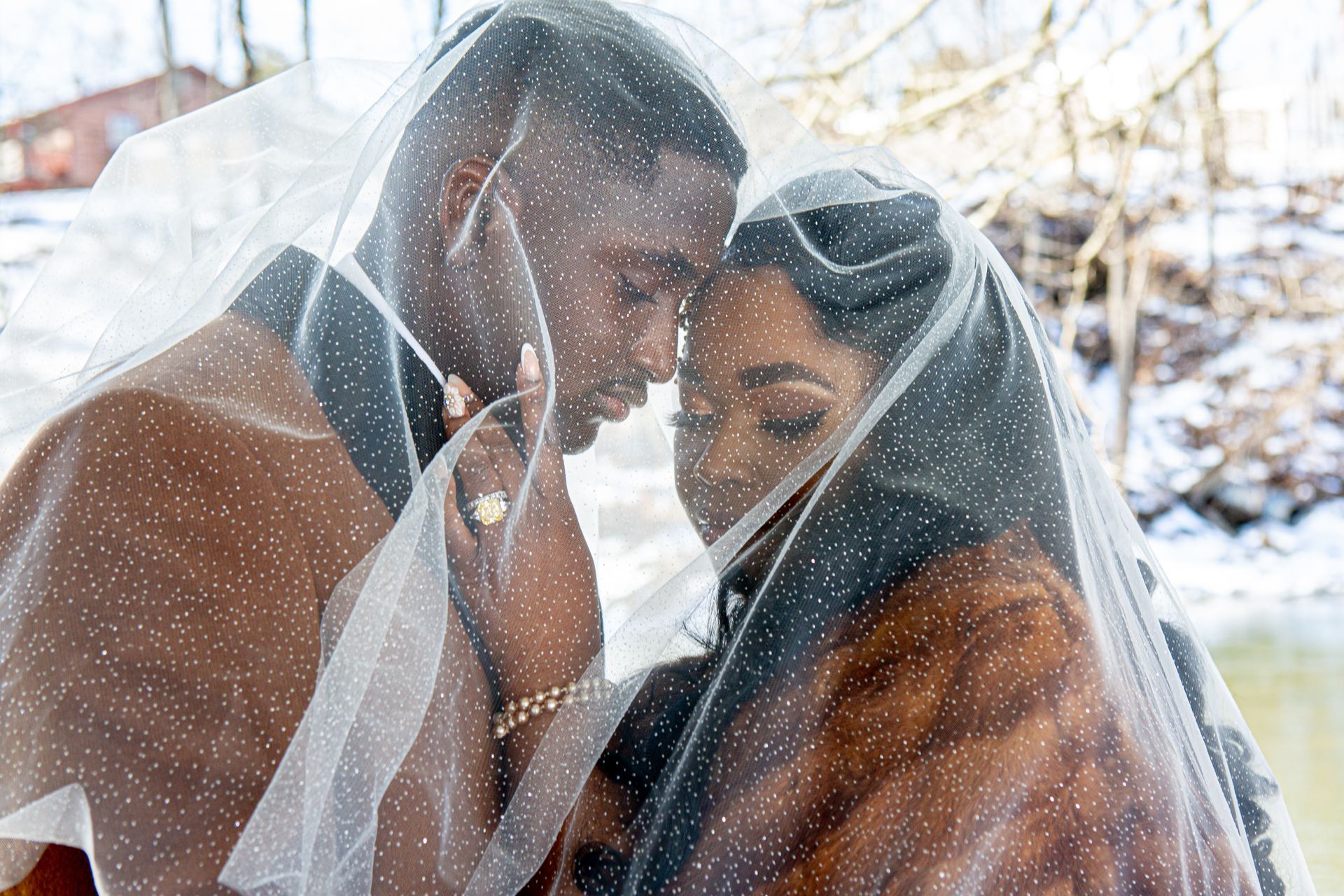 A couple faces each other intimately, heads bowed under a glittery, semi-sheer veil against a snowy, outdoor background.