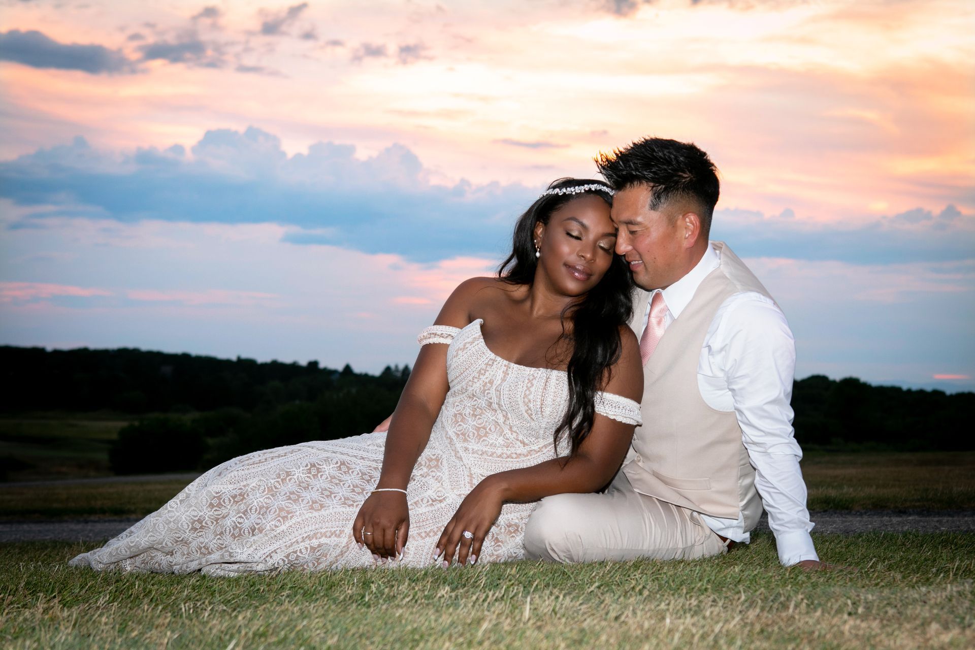 A couple sits in the grass at sunset, leaning heads together affectionately while dressed in formal wedding attire.