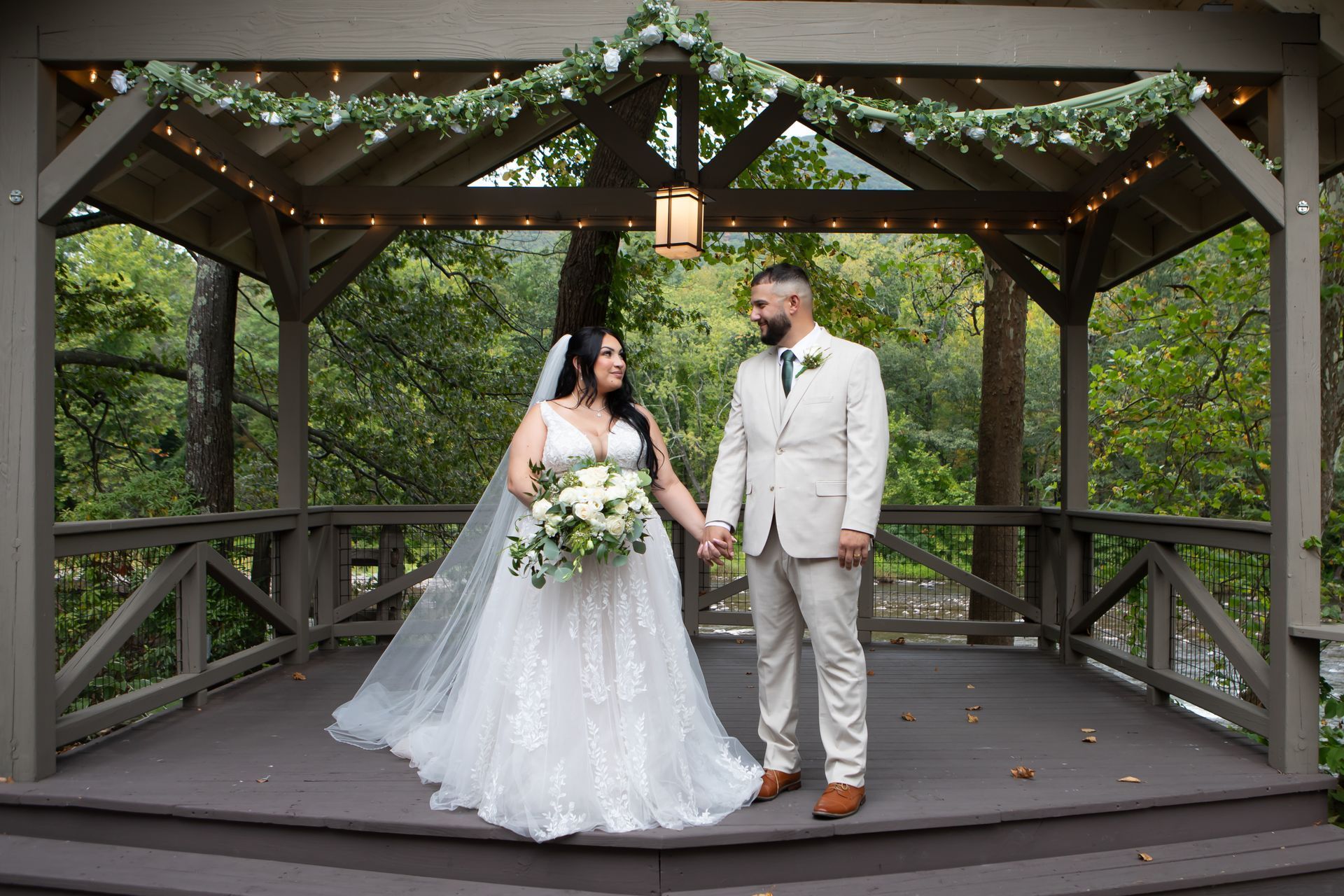 A wedding couple holds hands, smiling at each other inside a wooden gazebo decorated with greenery and fairy lights.