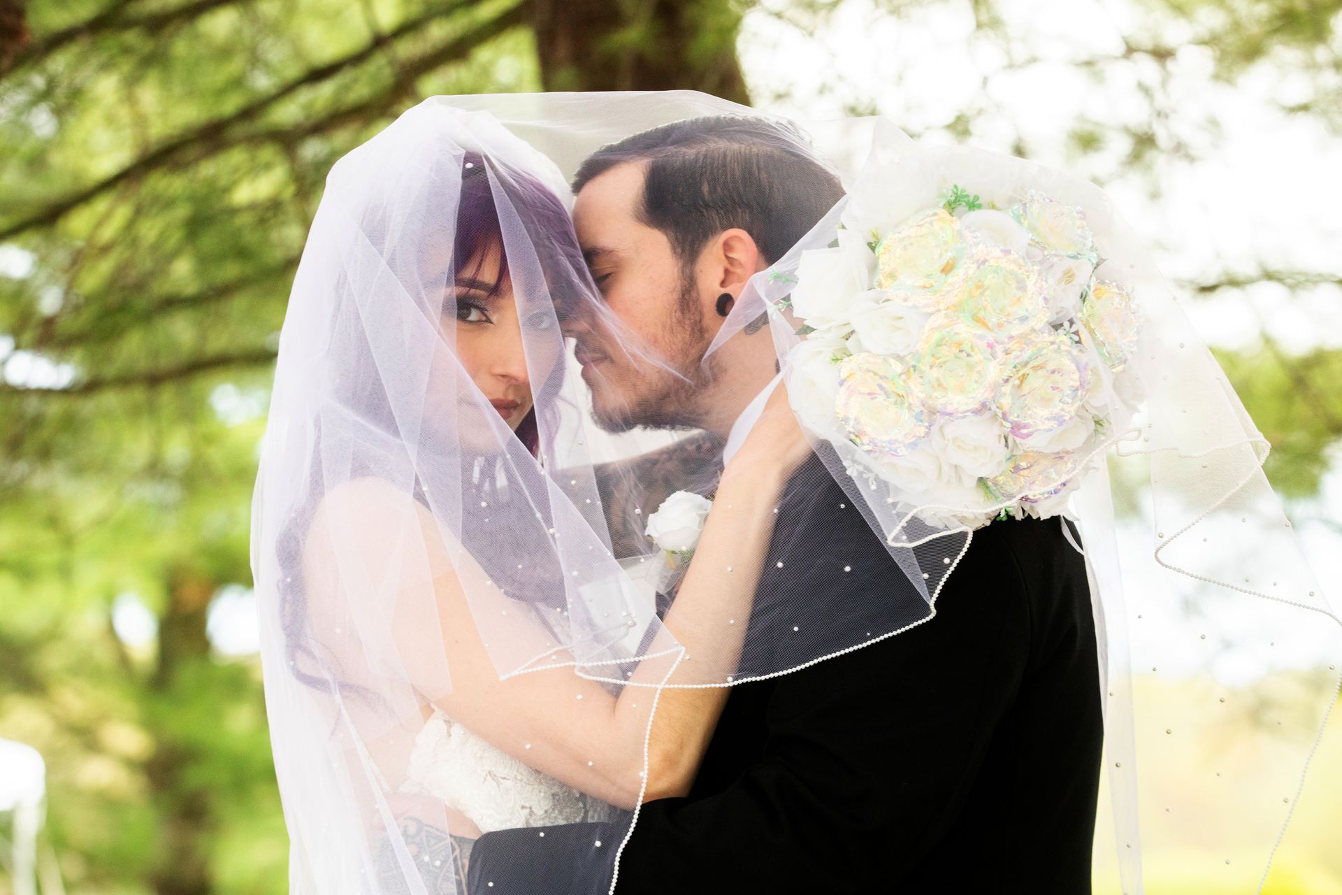 A bride and groom in a dark suit pose closely outdoors, sharing an intimate moment under a delicate lace bridal veil.