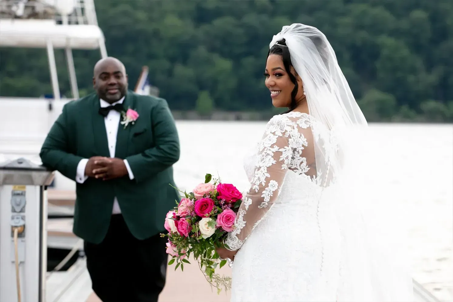 Bride in white dress looks back at groom in green suit, holding a bouquet, on a dock.