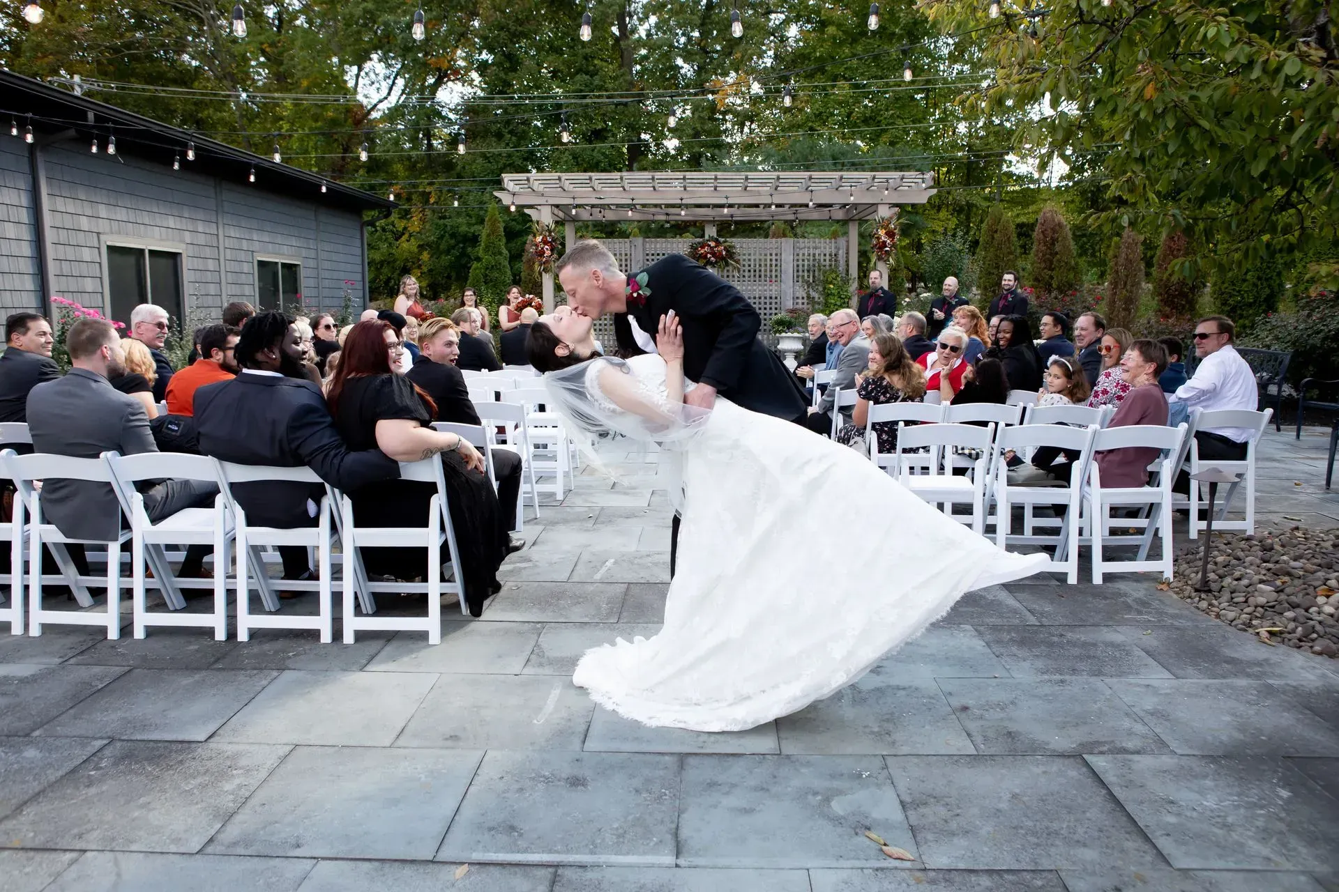 Newly married couple kissing, bride's dress flowing, guests watching at an outdoor wedding ceremony.