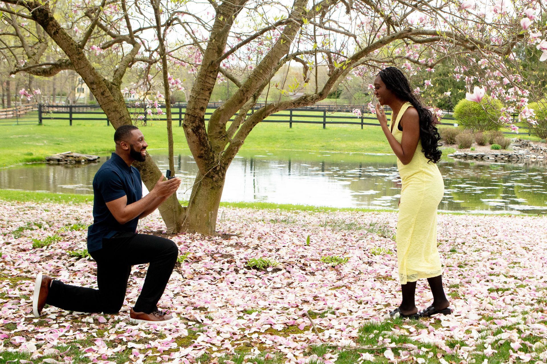 Man proposing on one knee to a woman in a yellow dress under blooming trees by a lake