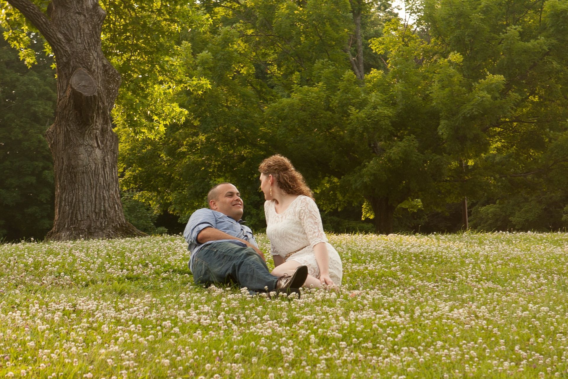 Couple sitting in a grassy flower field under trees, facing each other and smiling
