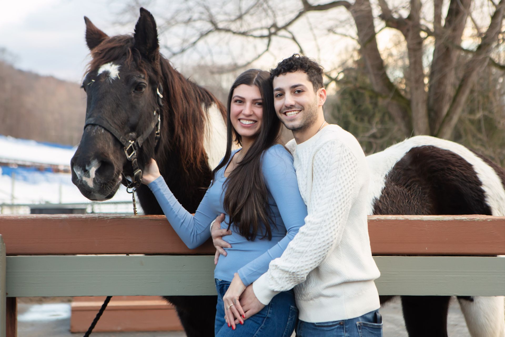 Couple standing beside a black-and-white horse by a wooden fence outdoors
