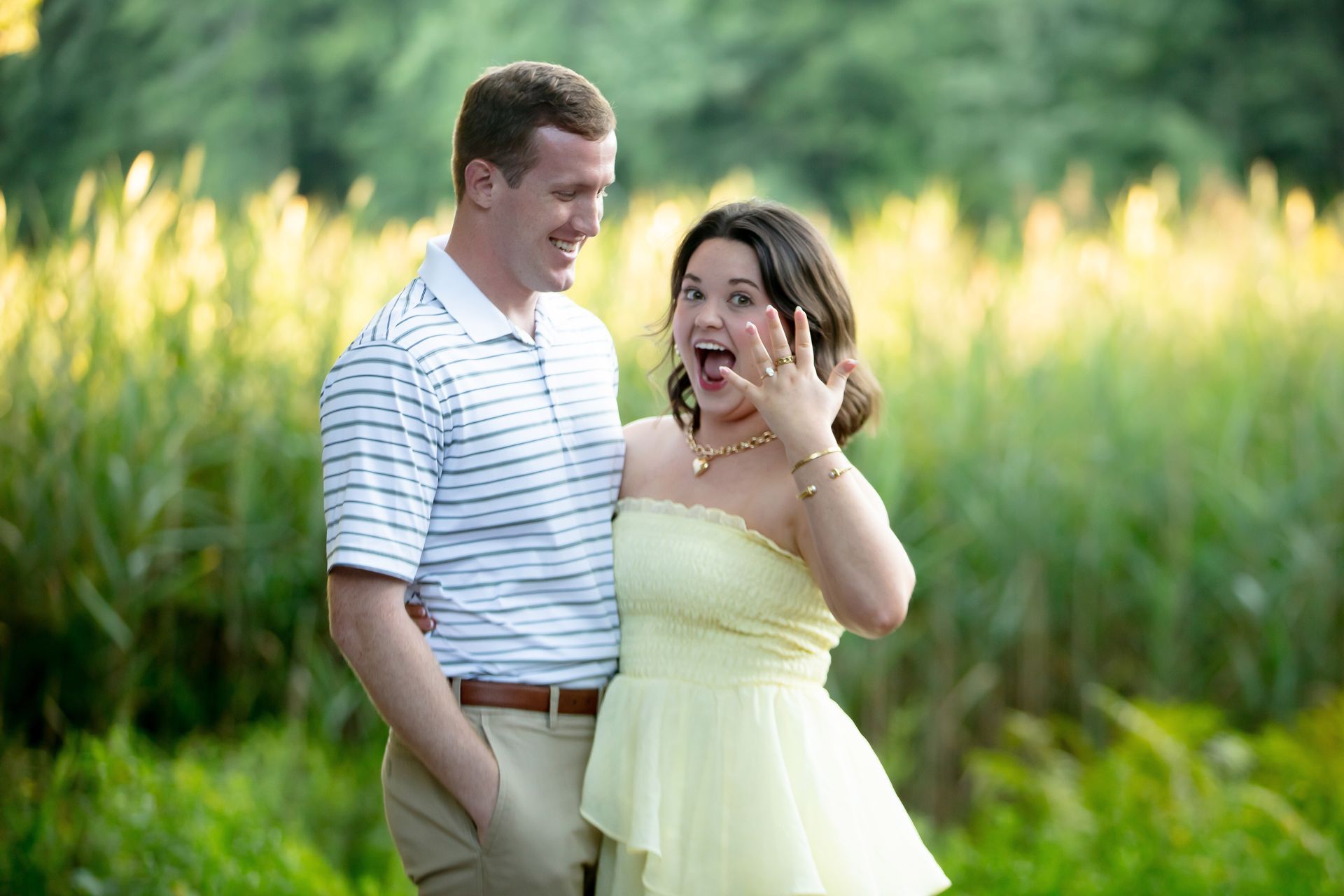Couple standing in a sunlit field, smiling and laughing, with green plants and tall grass behind them.