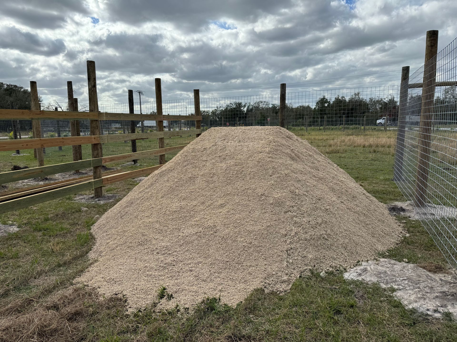 A pile of gravel is sitting in the middle of a field next to a chain link fence.