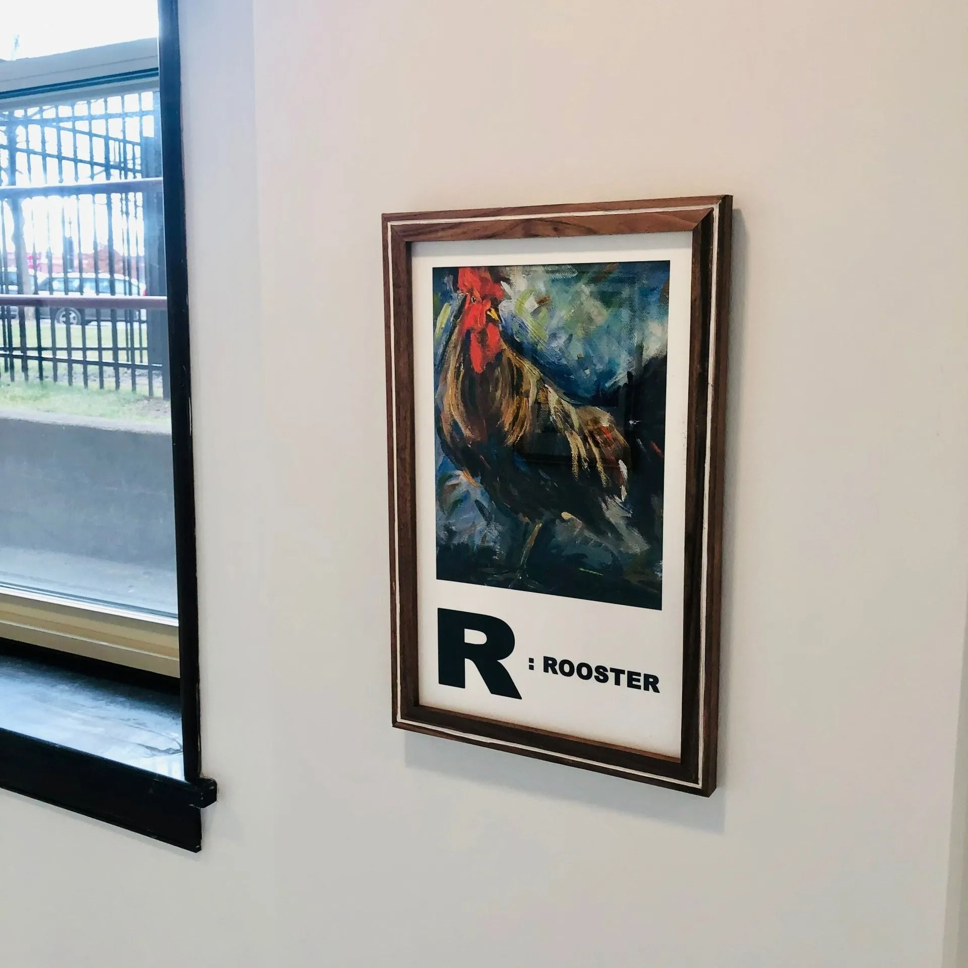 Framed rooster poster on a white wall beside a window