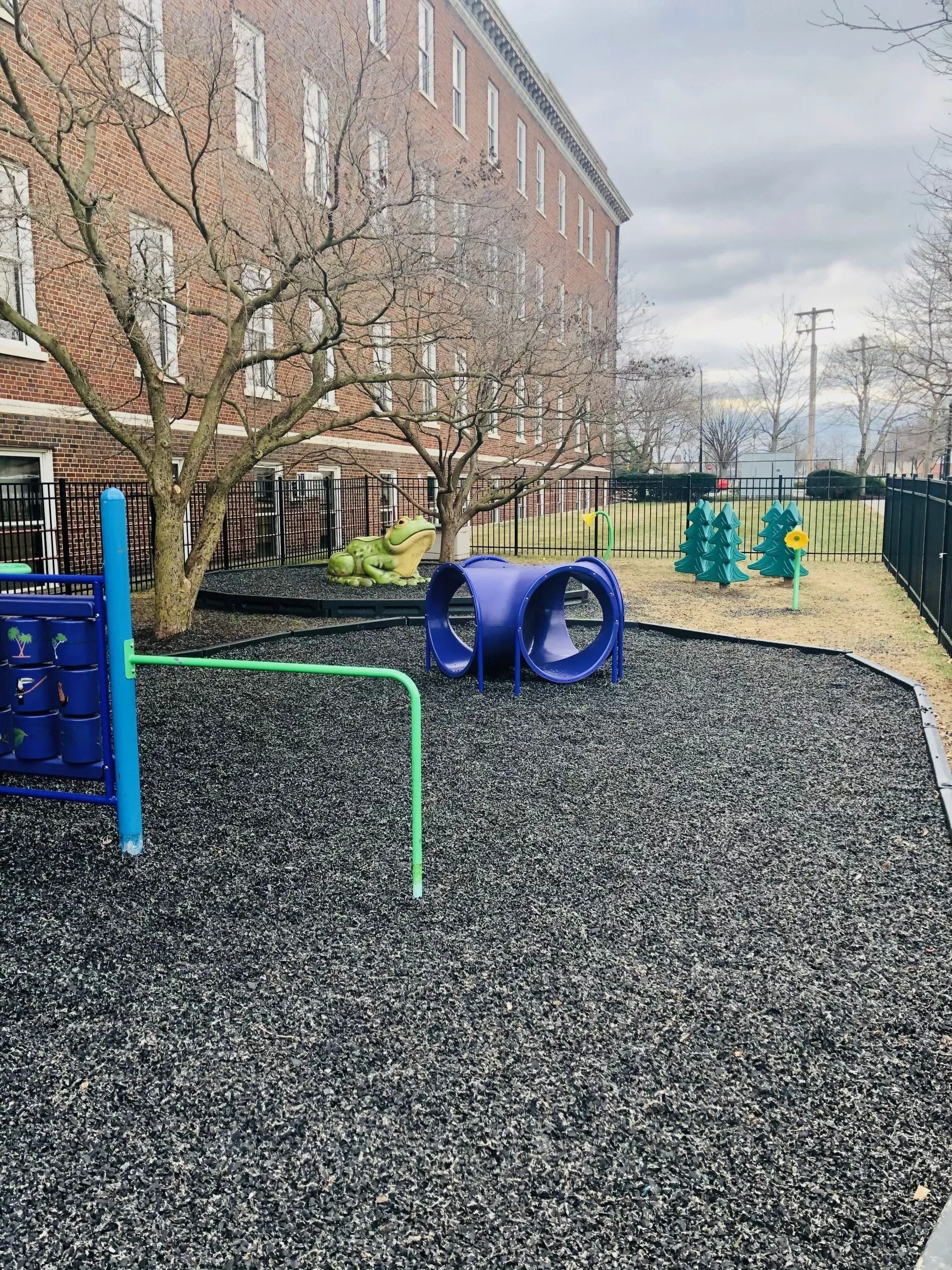Playground with blue tunnel climber and green bars on black rubber ground beside a brick building