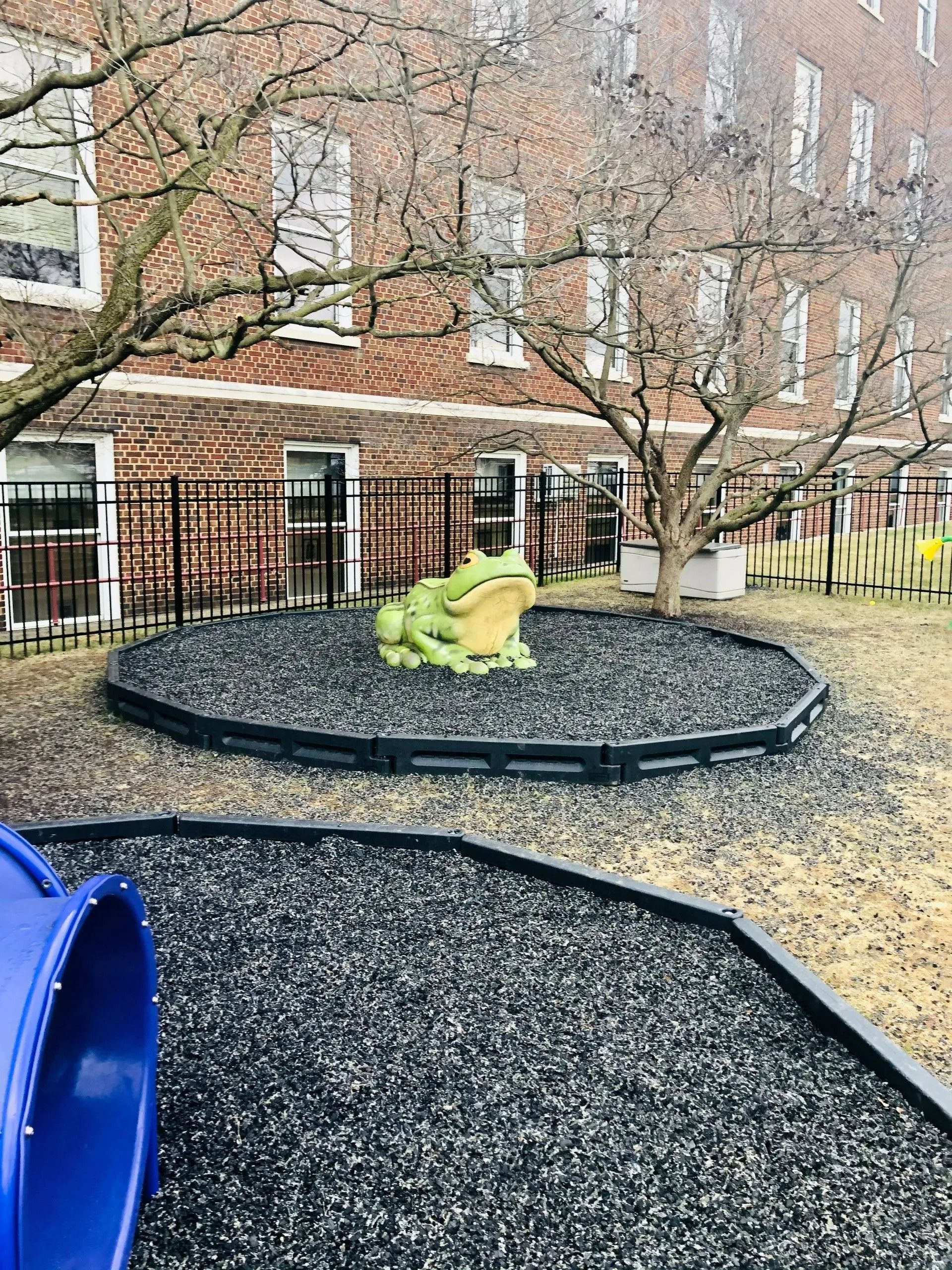 Bright green frog sculpture on a black gravel playground near a brick building and trees