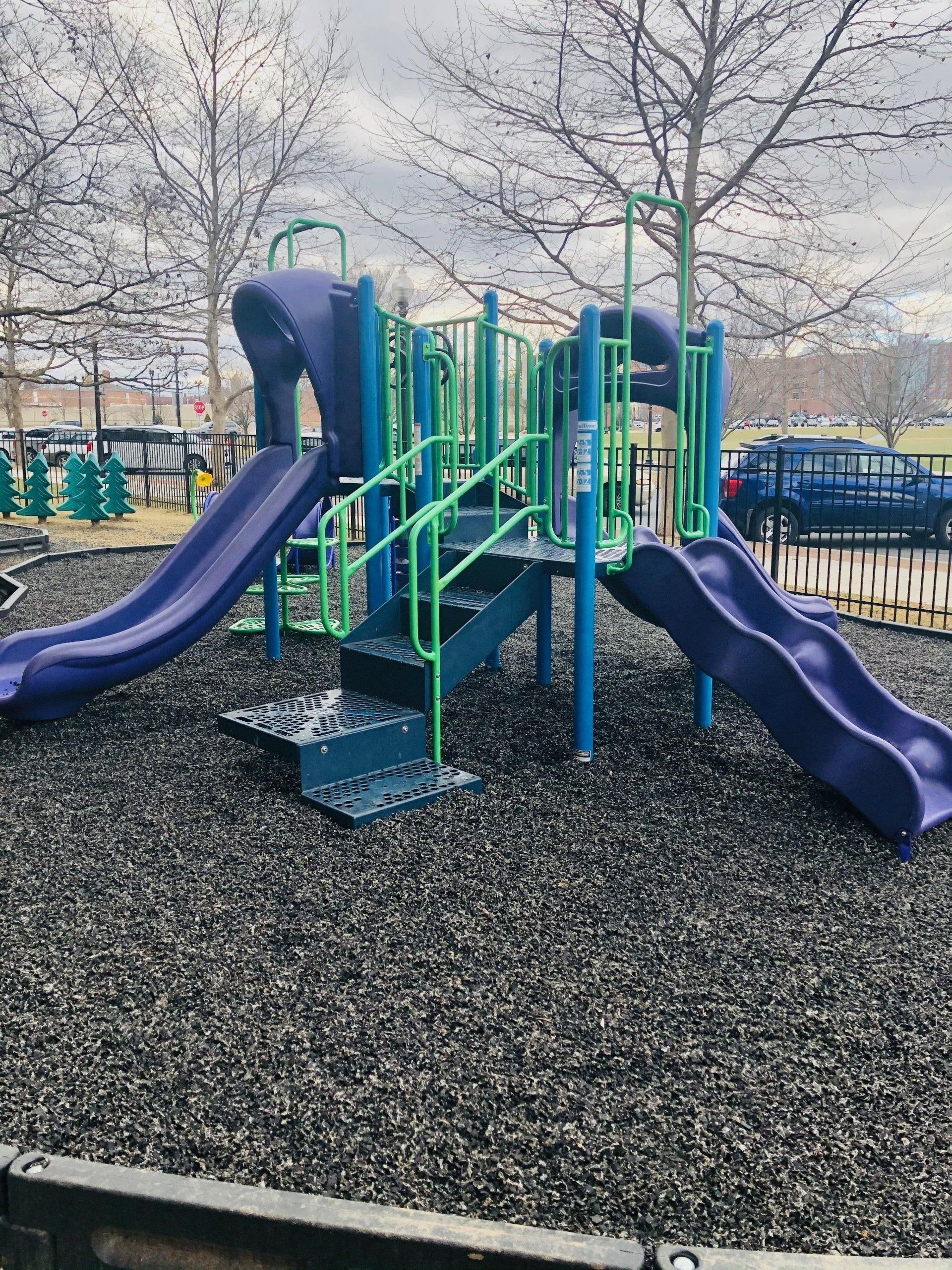 Outdoor playground with purple slides, green climbing bars, and leafless trees on a woodchip surface