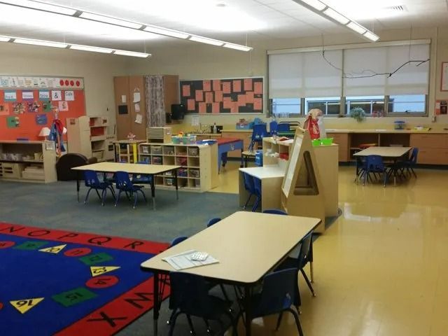 Empty classroom with desks, colorful rug, cubbies, and bright windows.