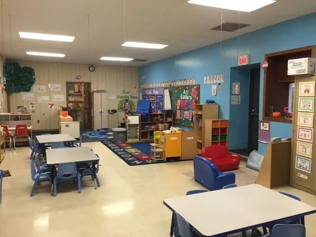 Bright preschool classroom with tables, shelves, colorful rugs, and play area under fluorescent lights