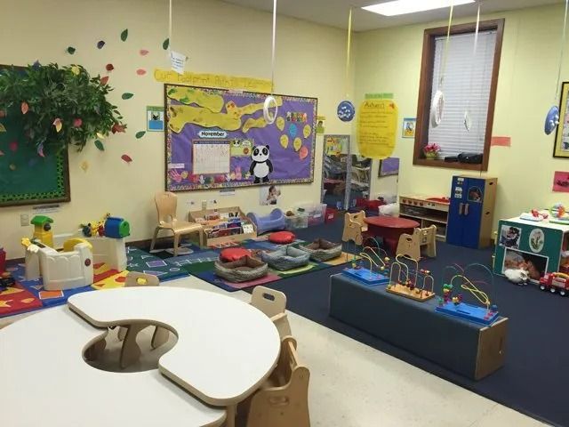 Colorful preschool classroom with tables, toys, bulletin boards, and play areas on a blue carpet