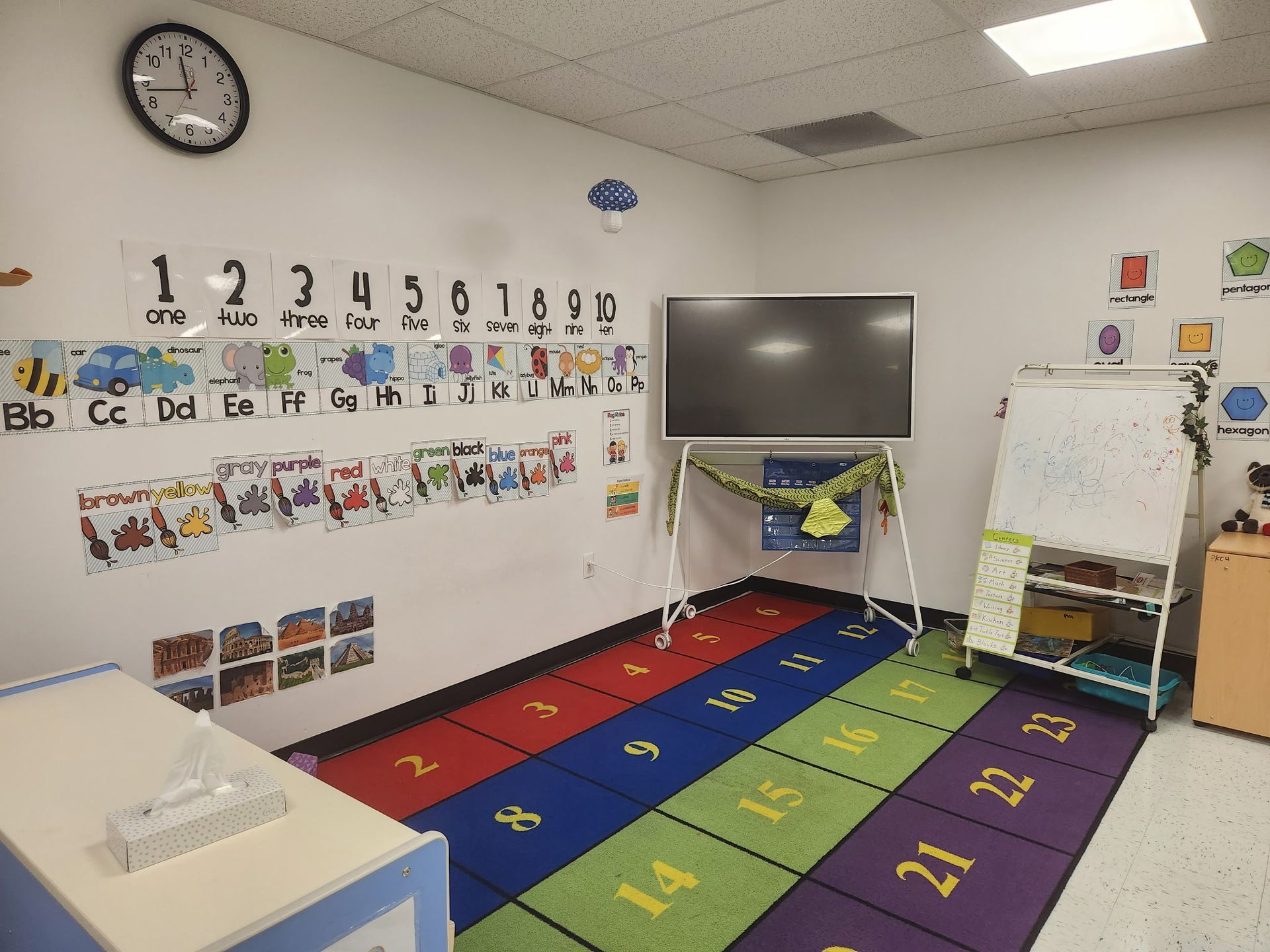 Bright preschool classroom with alphabet wall, colorful rug, TV, easel, and whiteboard
