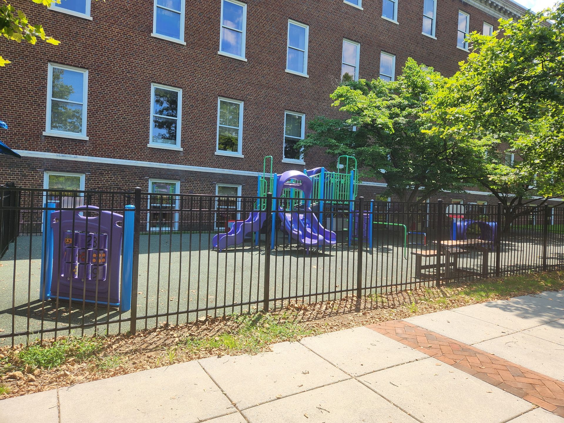 Small playground with purple climbing equipment behind a black fence beside a brick building