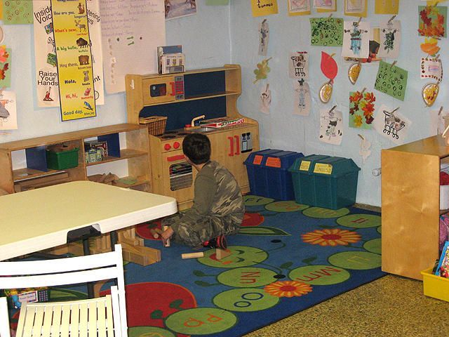 Child sitting on a colorful classroom rug facing shelves and wall art in a play area