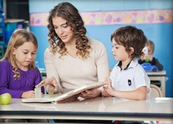 Teacher reading with two children at a classroom table, with a yellow ball nearby.
