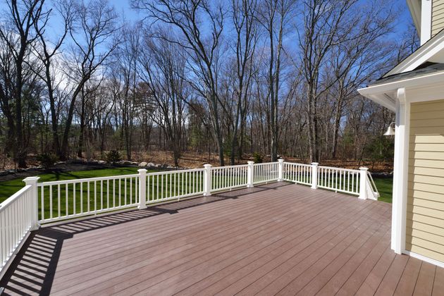 Wooden deck with white railing beside a house, overlooking trees on a sunny day