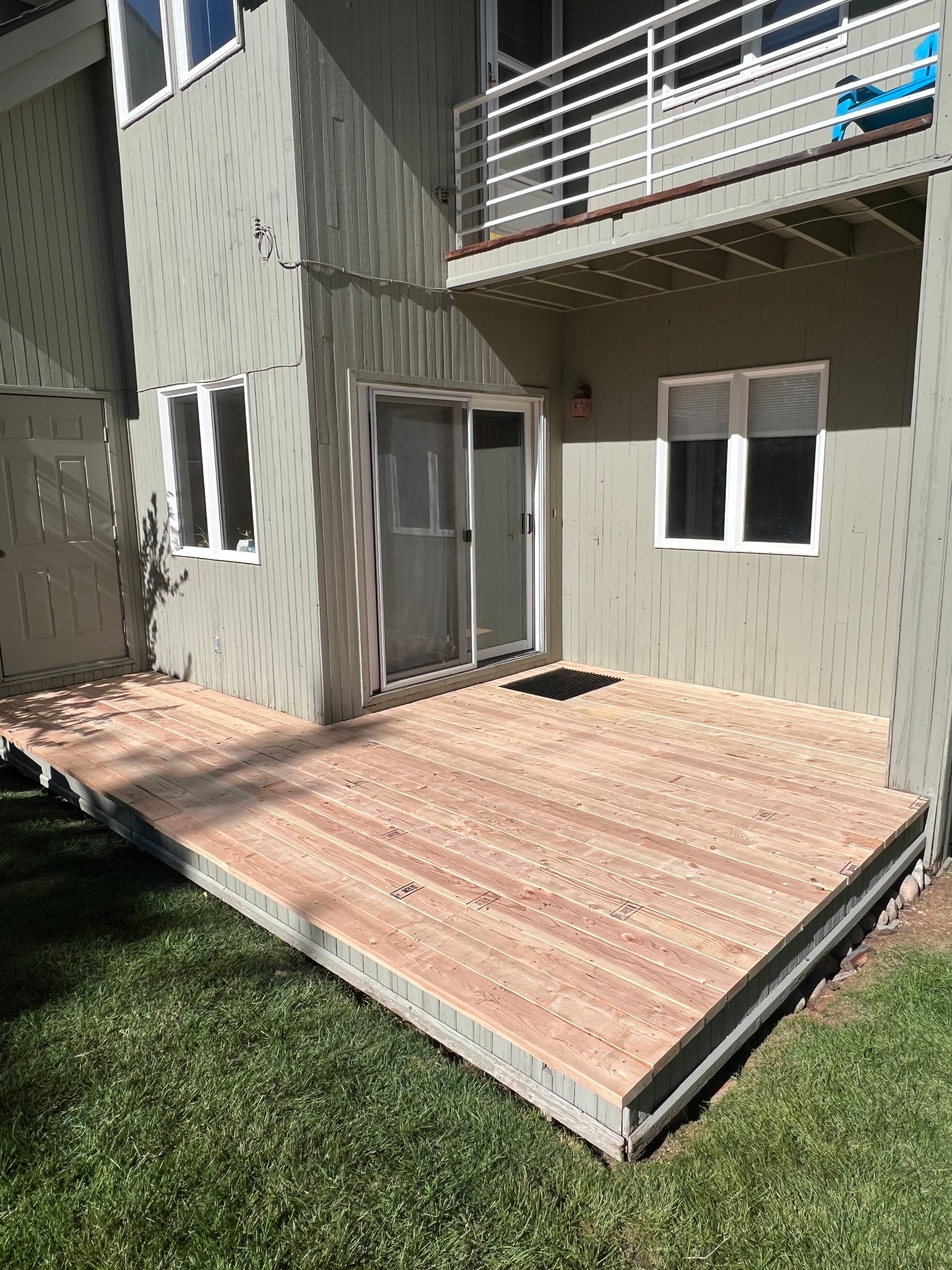 Backyard wooden deck and step leading to a gray apartment patio door with balcony above
