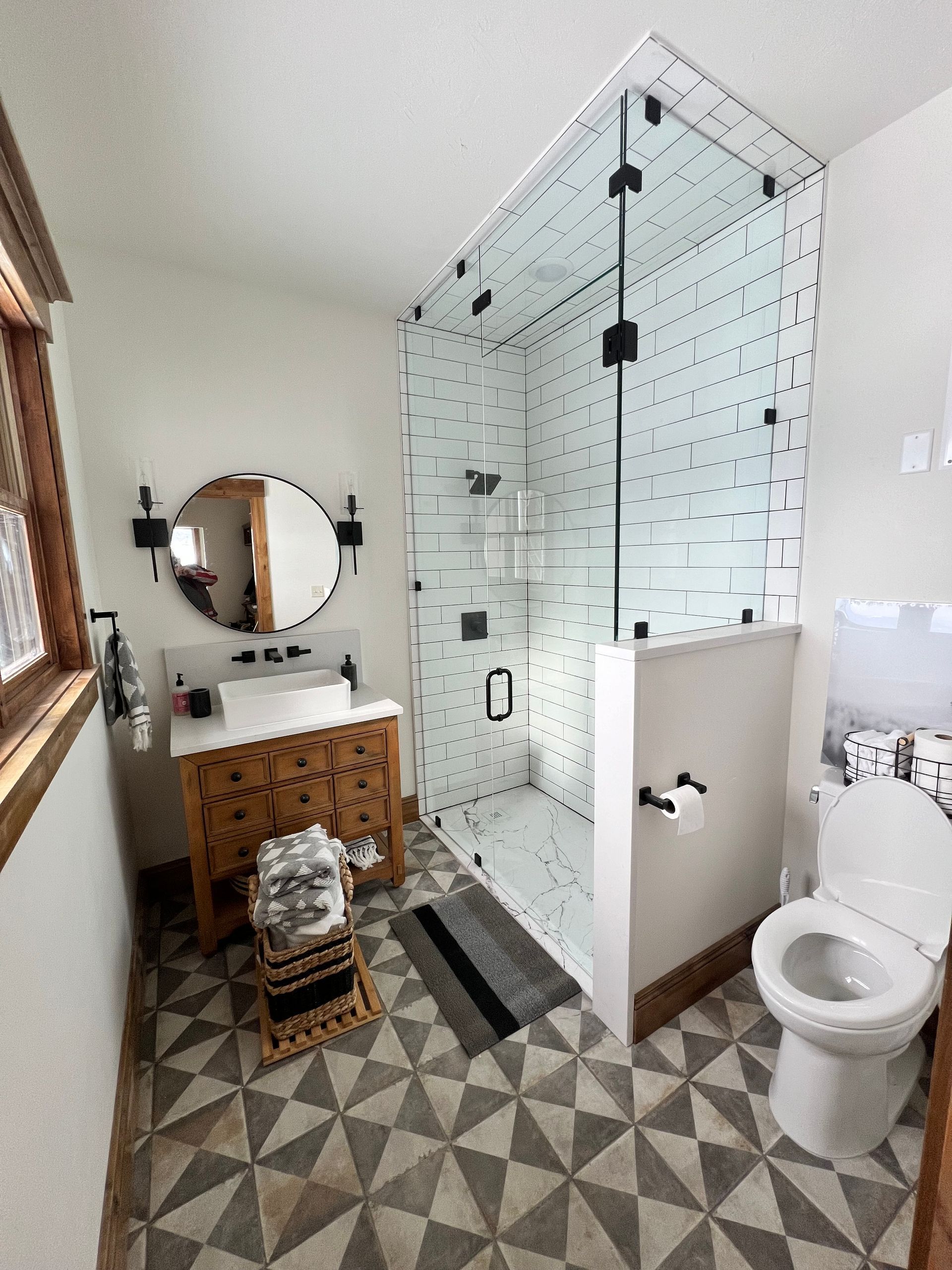 Modern bathroom with glass shower, white toilet, patterned tile floor, and wood vanity with round mirror