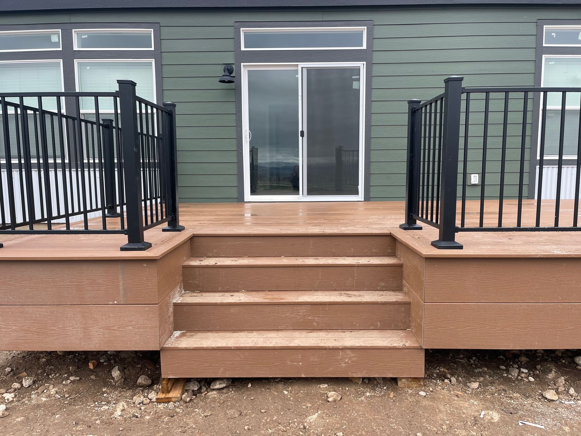 Wooden deck with black railings and steps leading to sliding glass doors on a green house exterior