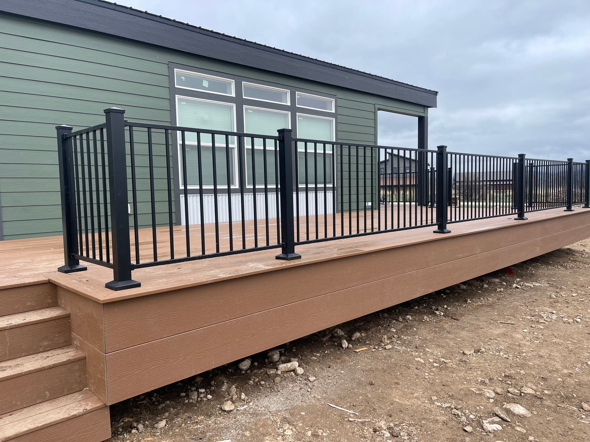 Raised wooden deck with black metal railing beside a green building under cloudy sky